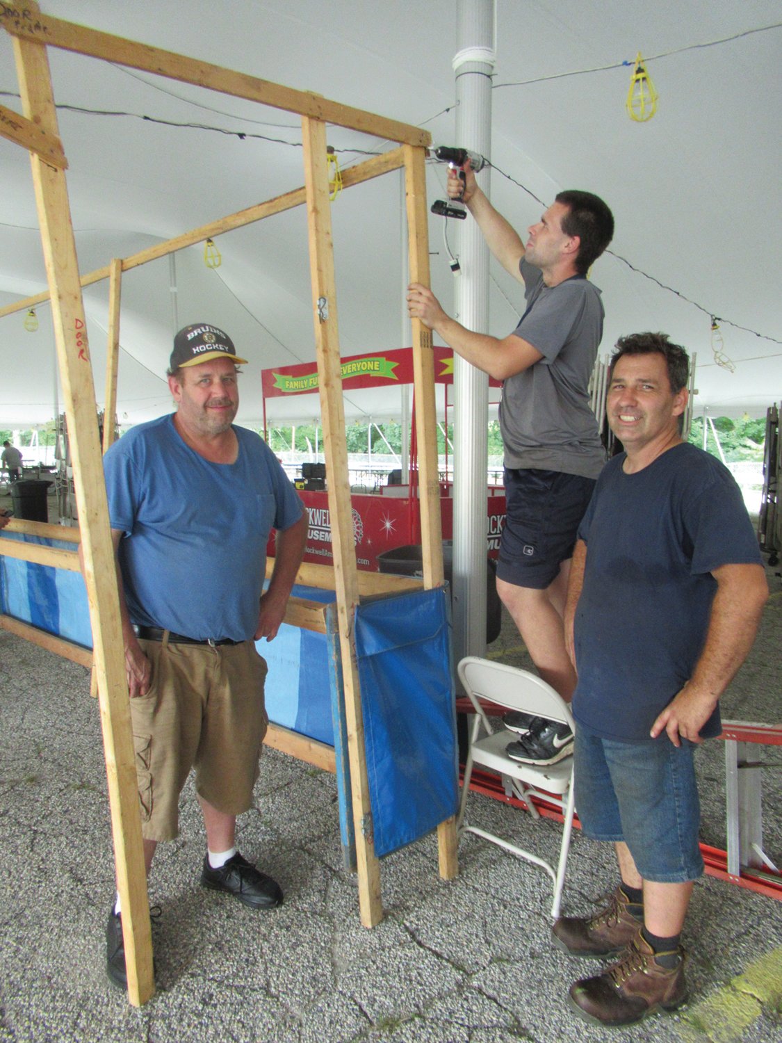 STRONG SUPPORT: Jeremy Bousquet nails a portion of the Women’s Guild Straw Game both as Bruce Baker and David Charron make sure the ladder doesn’t move Monday night while setting up under the “Big Top” for this weekend’s Saint Rocco’s Feast and Festival.